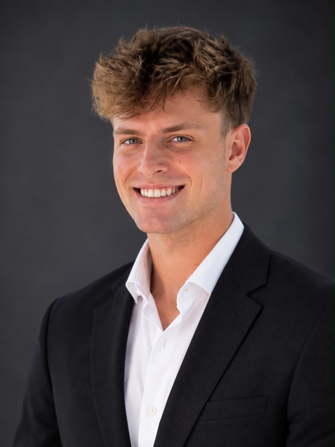 Young man smiling at camera wearing black blazer and white dress shirt against dark background